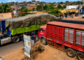 Pedestrians wait along the Kampala–Jinja Highway as Coca-Cola delivery trucks partially block the walkway, forcing road users to pause and navigate congestion caused by loading and offloading activities.