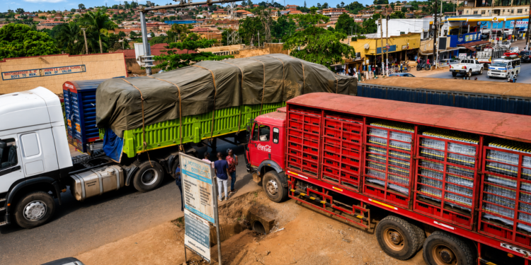 Pedestrians wait along the Kampala–Jinja Highway as Coca-Cola delivery trucks partially block the walkway, forcing road users to pause and navigate congestion caused by loading and offloading activities.