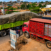 Pedestrians wait along the Kampala–Jinja Highway as Coca-Cola delivery trucks partially block the walkway, forcing road users to pause and navigate congestion caused by loading and offloading activities.